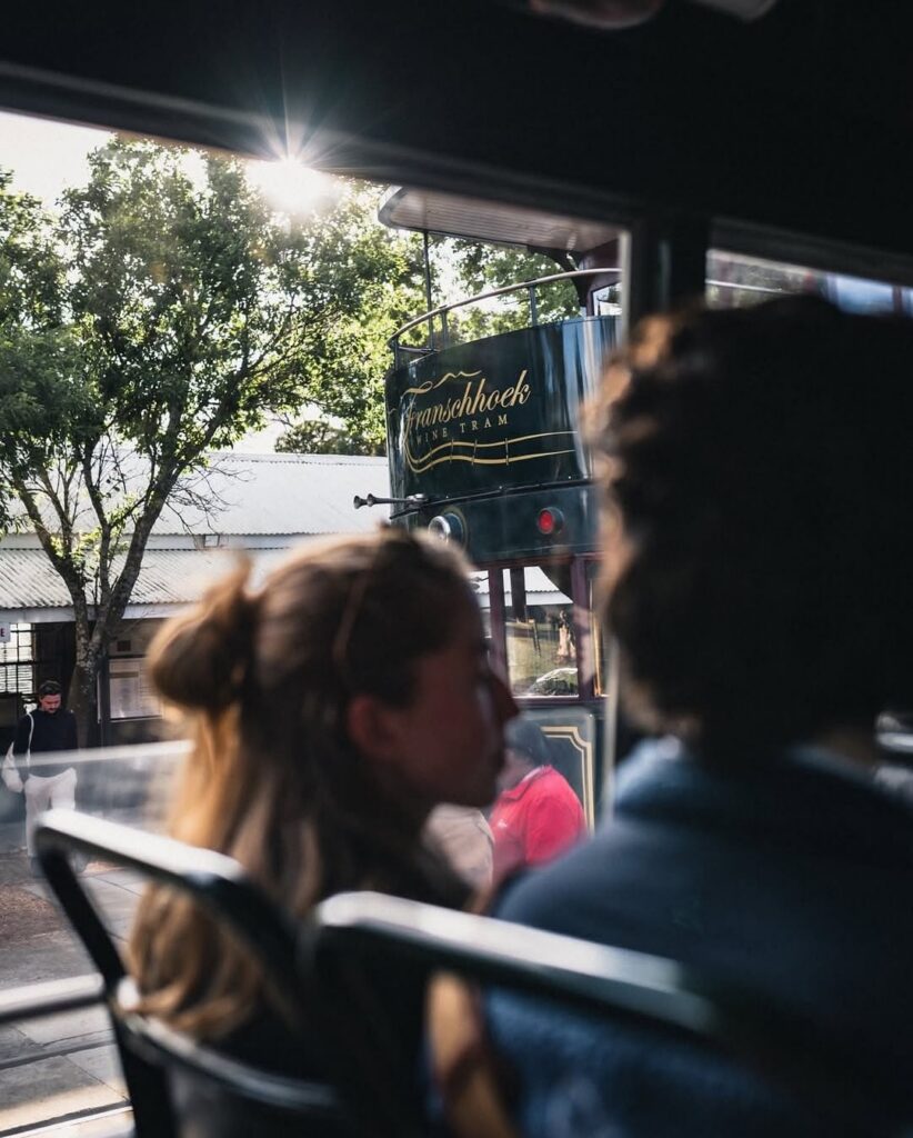 Passengers look out the window of a tram toward another Franschhoek Wine Tram on a sunny day.