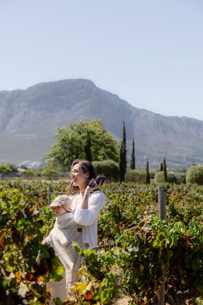 A smiling woman carries a baby in a sling while holding a bunch of grapes in a sunlit vineyard with mountains behind.